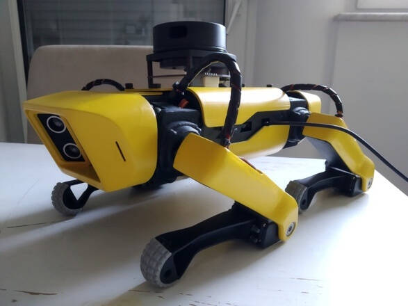 A yellow, four-legged robotic dog stands on a white table. The robot features a camera and sensors on its front and top, with black and yellow limbs and thick, textured feet. Cables and components are visible along its body.